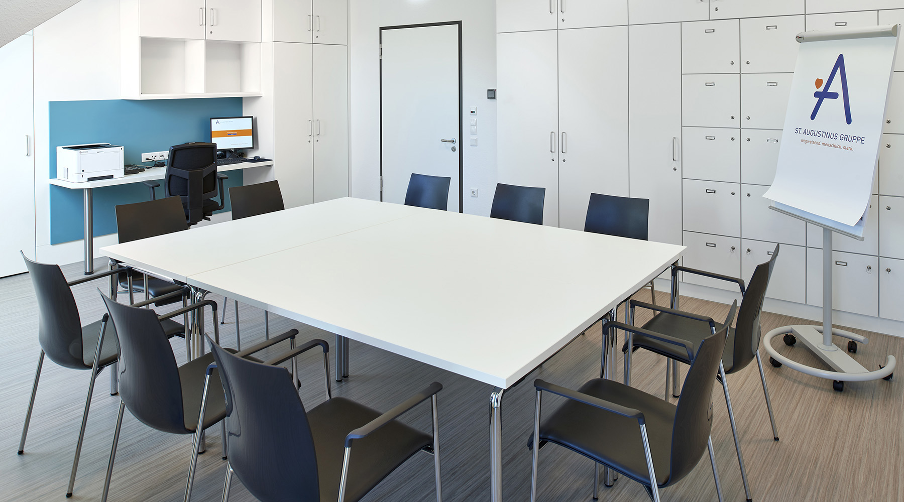 Ein Meetingraum mit einem großen weißen Tisch und blauen Stühlen drumherum, auf der rechten Seite steht ein Whiteboard.