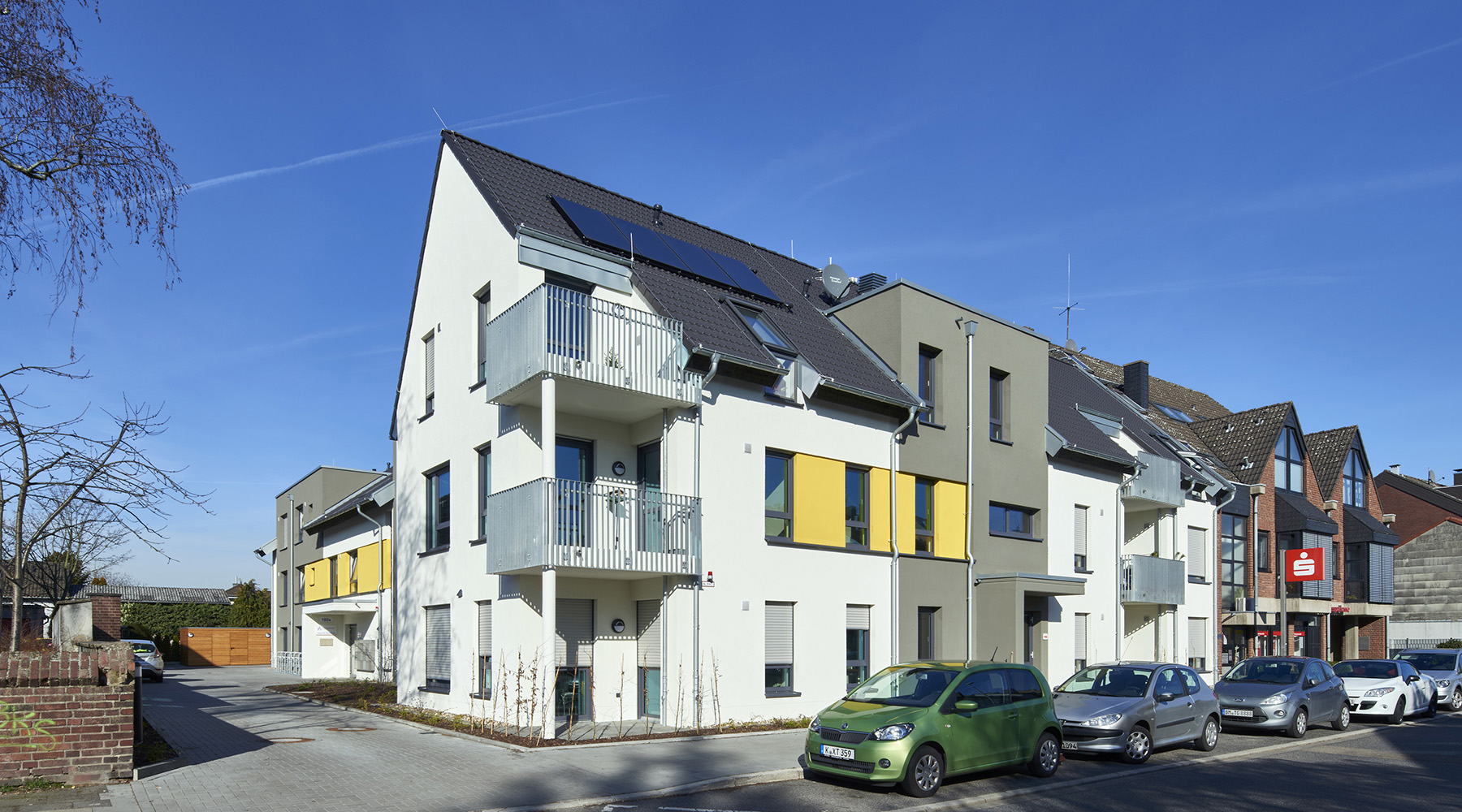 Ein modernes Gebäude mit Zeltdach und Balkonen. Es ist überwiegend weiß, mit grauen und gelben Elementen.