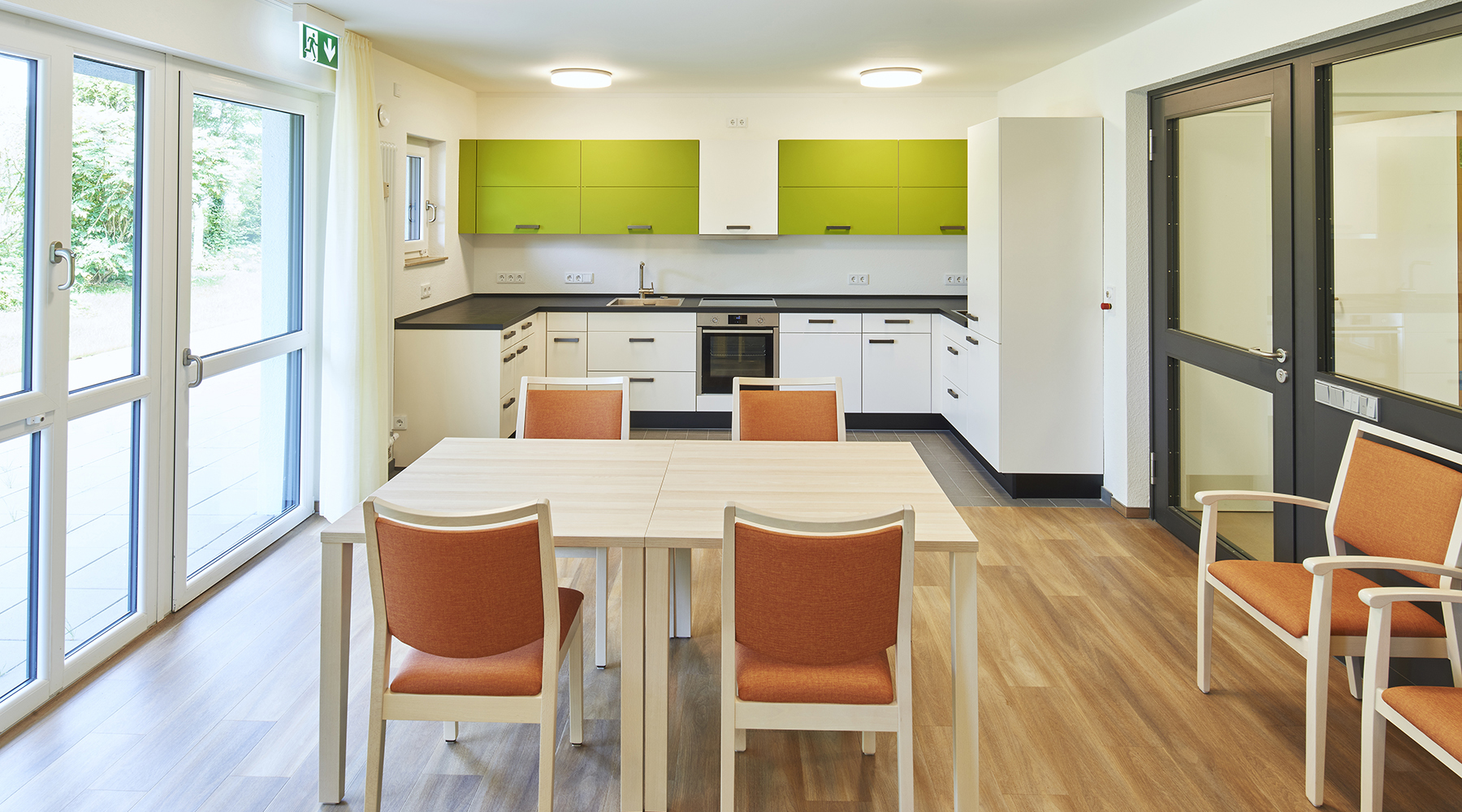Holztisch mit braun-bezogenen Stühlen steht in der Mitte des Raumes. Im Hintergrund ist eine weiße Küche mit schwarzer Arbeitsplatte und grünen Hängeschränken.