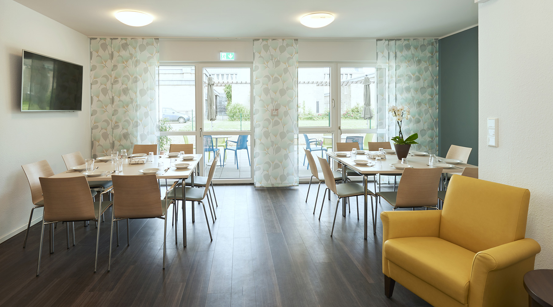 Ein Speisesaal mit direktem Zugang zur Terrasse. Zwei große gedeckte Esstische stehen im Raum und weiter vorne gelbe Sessel um einen Kaffeetisch.