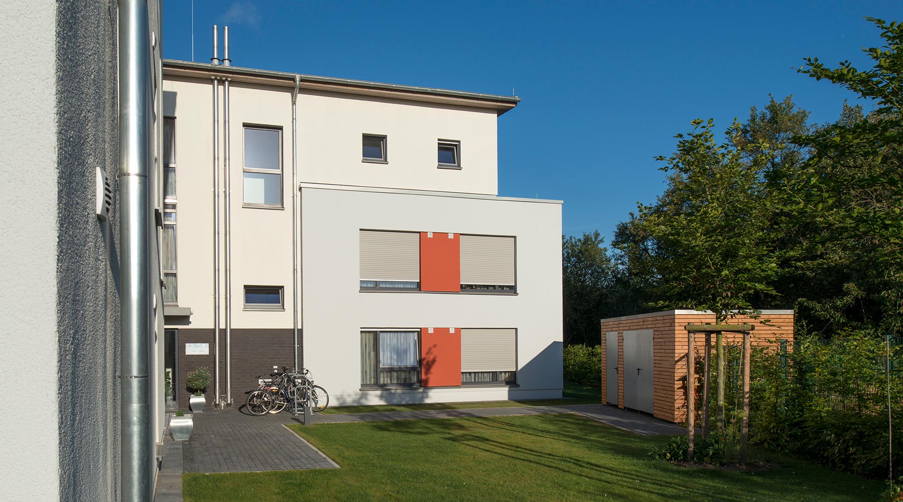 Stationäres Wohnheim Haus Matthias in Neuss. Ansicht vom Garten: Helle Fassade mit orangen Akzenten.